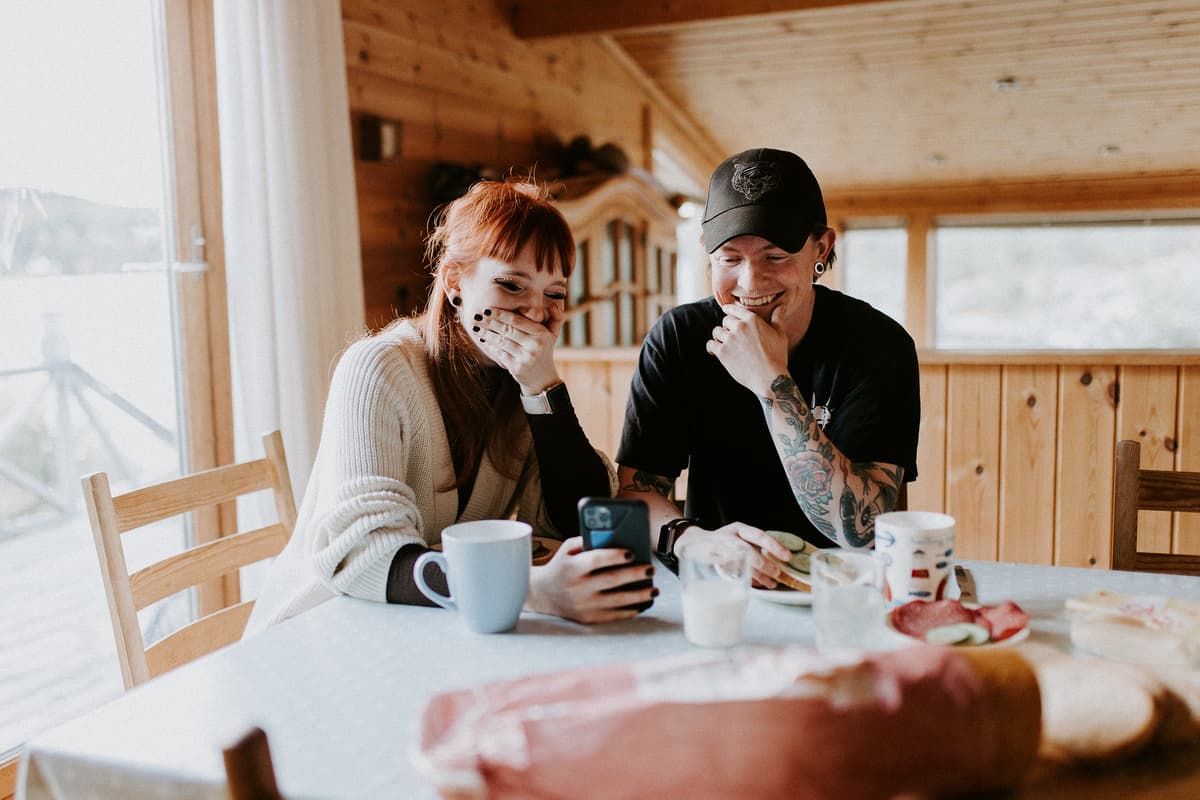 Foto: Line Owren Fotografi AS Bilde av en mann og en dame som sitter ved et bord. Damen holder en mobiltelefon i hånda og begge ser på skjermen og smiler.