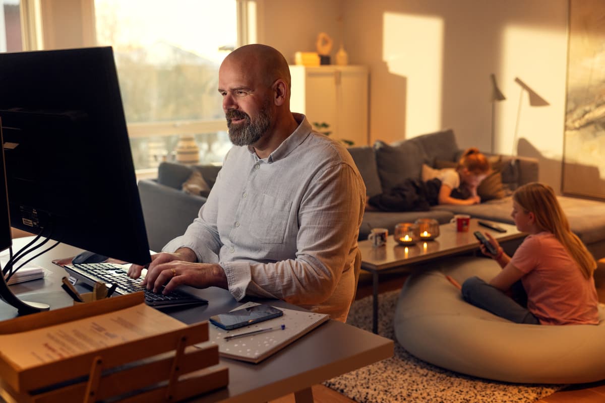 Mine Sider Altibox Mann sitter ved datamaskinen på stua med barn i bakgrunnen.