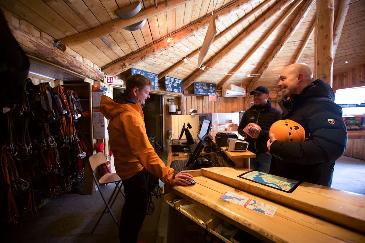 Kjøper billett på Rypetoppen En mann står bak kassa og skal selge billetter til to menn som holder hver sin klatrehjelm i hånda.
