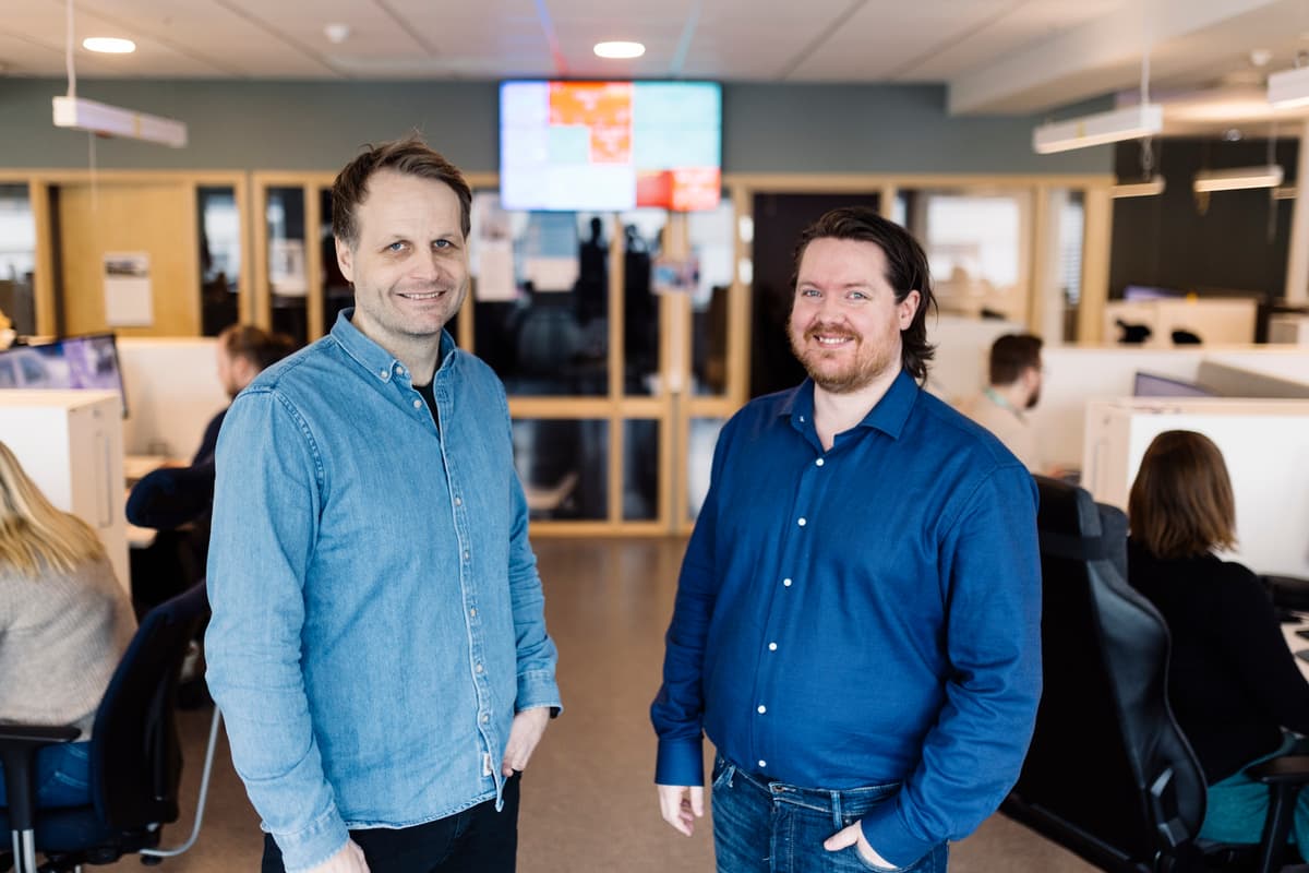 To menn står i et åpent kontorlandskap hvor vi ser ryggen på flere som sitter og jobber i bakgrunnen. To menn står i et åpent kontorlandskap hvor vi ser ryggen på flere som sitter og jobber i bakgrunnen.