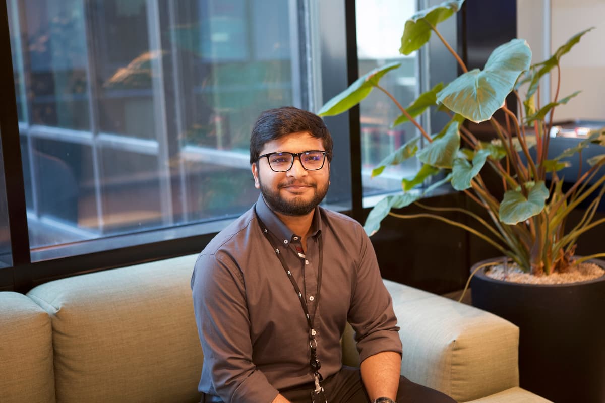 Smilende mann med briller sitter i en sofa som står ved siden av en stor grønnplante Smilende mann med briller sitter i en sofa som står ved siden av en stor grønnplante
