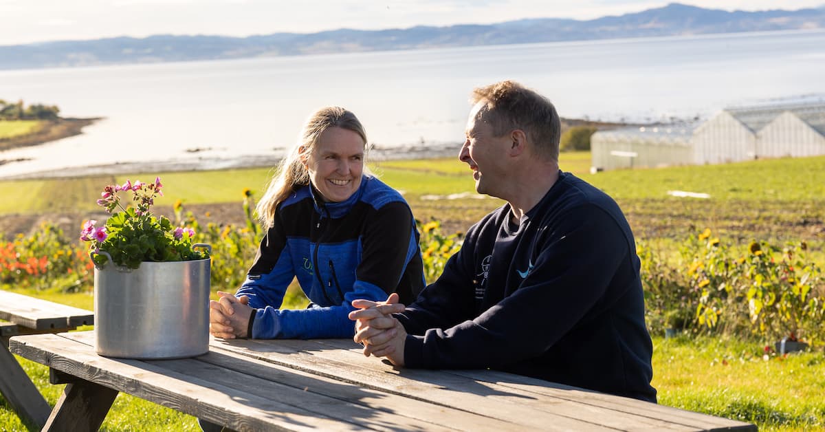 Utendørs Mann og dame sitter ved en benk med fjord og åkre bak seg